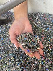 Een hand schept stukjes gerecycled plastic uit een grote bak.