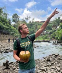 Rick wijst omhoog en staat voor een rivier in tropische omgeving, hij houdt een tropische vrucht vast,